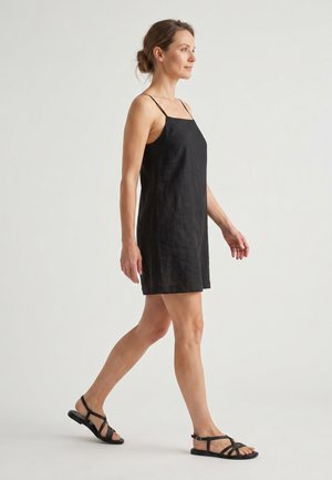 Kvinde går, iført en sort ærmeløs kjole med tynde stropper og sorte flade sandaler mod en ensfarvet lys baggrund.