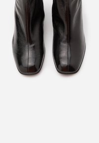 Botas de tobillo de cuero negro con acabado brillante, punta cuadrada y cremallera lateral. Presentan una textura suave y un diseño aerodinámico.