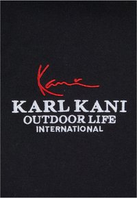Tela negra con texto blanco bordado "KARL KANI OUTDOOR LIFE INTERNATIONAL" y una firma roja, destacando el contraste y la marca.