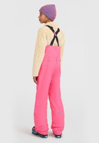 Felle roze sneeuwoverall met zwarte verstelbare banden, gecombineerd met een crème fleecejack en een veelkleurige gestreepte muts.