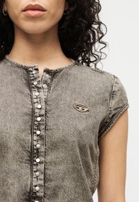 Κοντινό πλάνο ενός ατόμου που φοράει μια ξεθωριασμένη γκρι πουκαμίσα με κουμπιά, κοντά μανίκια και ένα οβάλ κεντημένο λογότυπο στο στήθος.