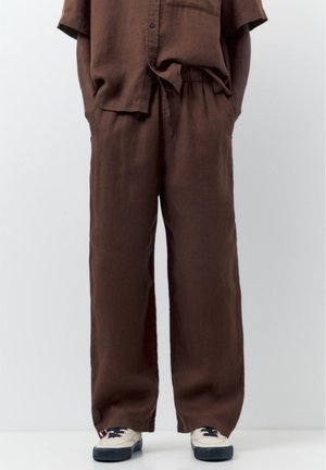 Persoană purtând pantaloni largi maro din in și cămașă cu mâneci scurte asortată, mâinile în buzunare, asortați cu adidași albi cu tălpi negre.