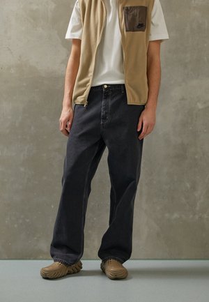 Persoon draagt een beige vest met rits over een wit T-shirt, losse zwarte jeans en beige instappers, staand tegen een grijze textuurmuur.