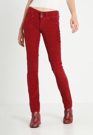 Femme portant un pantalon en velours côtelé rouge ajusté avec fermeture à double bouton et des bottines rouge à motif peau de serpent, debout devant un fond blanc.
