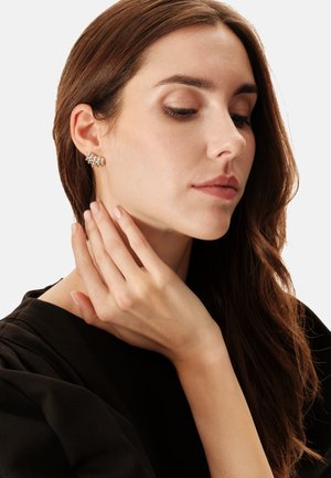 Pendientes de plata con diseño ondulado y detalles texturizados, llevados en la oreja de una modelo. Su mano descansa sobre el cuello, mostrando uñas naturales.