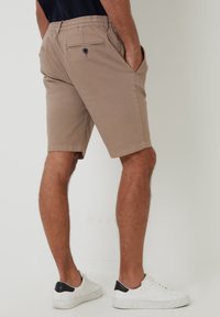 Shorts kaki avec une ceinture élastique, deux poches latérales et une poche arrière avec fermeture à bouton. Portés avec des baskets blanches.