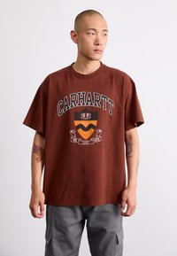 Brązowa bawełniana koszulka z grafiką z tekstem "CARHARTT" oraz wzorem tarczy. Krótkie rękawy, luźny krój i okrągły dekolt.