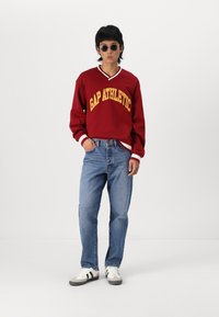 Red sweatshirt with "GAP ATHLETIC" in yellow lettering, paired with blue jeans and white sneakers featuring black stripes. Casual look.