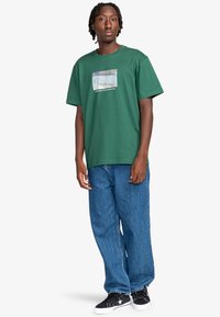 Grøn bomulds t-shirt med en firkantet grafik, der har teksten "Hilsner fra Barcelona". Casual fit med korte ærmer, parret med blå jeans.