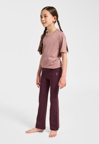 Dziewczyna z warkoczami stoi boso, ubrana w liliową koszulkę i ciemnofioletowe spodnie na jednolitym jasnym tle.