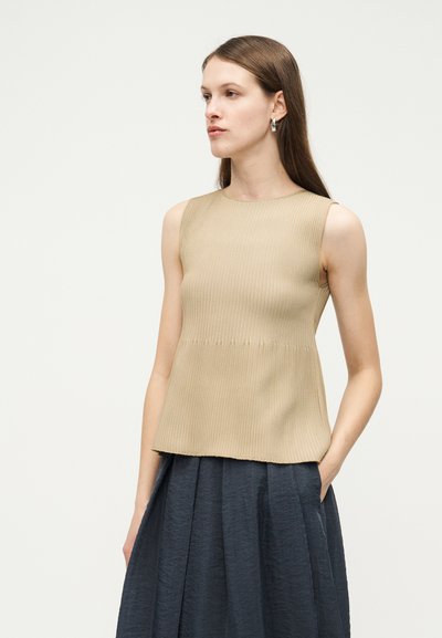 Femme portant un haut beige sans manches côtelé et une jupe taille haute bleu foncé, regardant sur le côté avec une main dans la poche de la jupe.