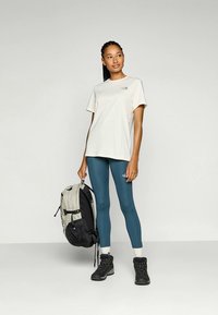 Mujer de pie, vistiendo una camiseta blanca, leggings azules y botas de senderismo, sosteniendo una mochila gris y negra frente a un fondo sencillo.