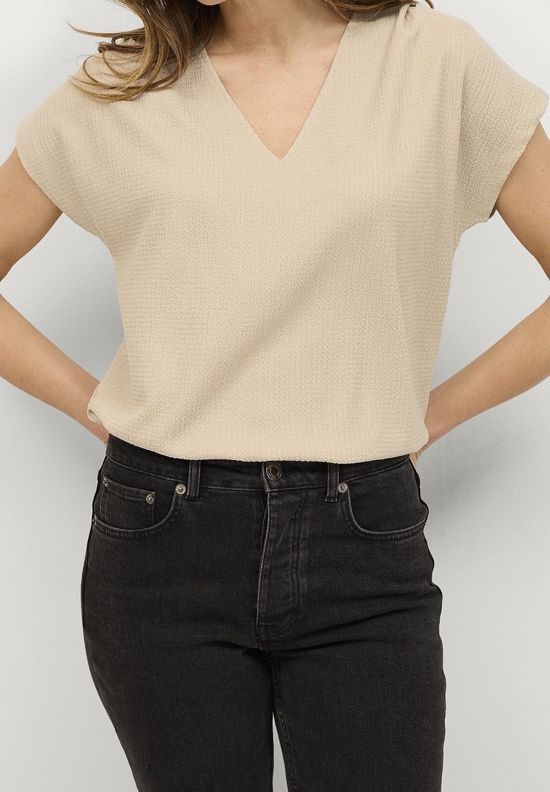 Haut beige texturé à manches courtes avec un col en V, associé à un jean noir taille haute doté de poches avant et de détails subtils en métal.