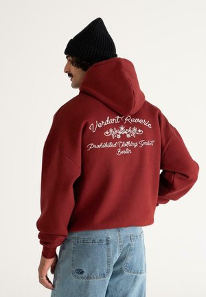Man wearing maroon hoodie with "Verdant Reverie, Prohibited Clothing GmbH Berlin" text and floral design on back, black beanie, and light blue jeans.