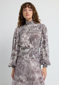 Bluse med blomsterprint i lys lilla og hvid, med høj hals, smocket talje og ballonærmer med elastiske manchetter. Glat tekstur.