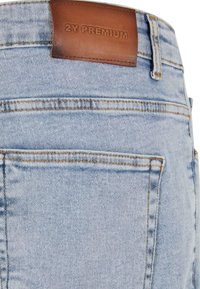 Vaqueros de mezclilla azul claro con un acabado texturizado, que presentan un parche de cinturón de cuero marrón grabado con "2Y PREMIUM". Diseño de cinco bolsillos.