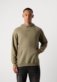 Jongeman in een olijfgroene Marc O'Polo Denim hoodie met een voorzak en zwarte spijkerbroek, staand tegen een effen witte achtergrond.