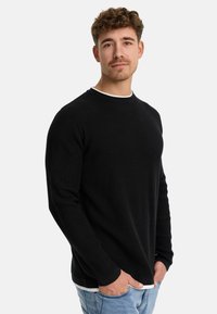 Crna longsleeve majica sa teksturiranim pletenjem, okruglim izrezom i bijelim naglaskom na ovratniku. Nosena preko svijetloplavih traperica.