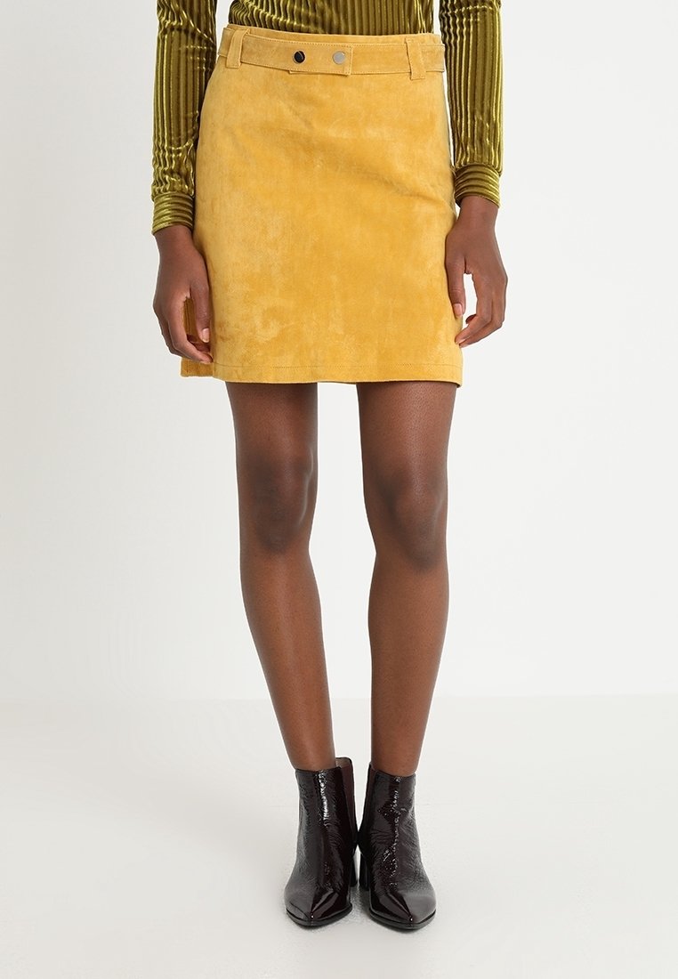 Mini-jupe en daim jaune avec un devant plat, des passants de ceinture et deux poches avant, associée à des bottines noires brillantes.