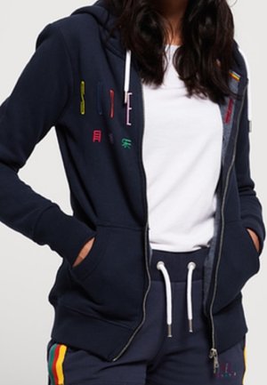 Woman wearing navy blue zip-up hoodie with colorful lettering and matching navy sweatpants with rainbow stripes and white drawstrings.