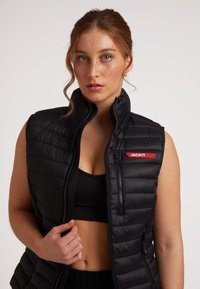 Gilet imbottito nero con design a righe verticali, colletto alto e etichetta rossa a contrasto "JACKIT". Presenta due tasche con zip.