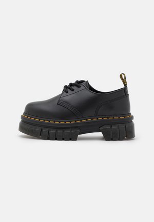 Zapato de piel negro con cordones, suela gruesa con ranuras y costuras amarillas alrededor del borde de la suela, mostrado de perfil lateral.