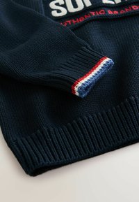 Maglione lavorato a maglia di colore blu navy con polsini a coste. Presenta righe bianche, rosse e blu sui polsini. Grande logo bianco e rosso sul davanti.