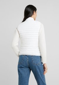 White puffer vest with a ribbed knit sweater portion, featuring a stand-up collar and horizontal padding design. Worn with blue jeans.