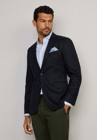 Blazer marine avec des revers crantés, deux boutons et poches à rabat, porté sur une chemise bleu clair avec un pochette à motifs et un pantalon vert.