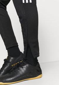 Černé sportovní kalhoty s bílými třemi pruhy, se zipem na kotníku a strukturovanou tkaninou. Spárované s černými sportovními botami.