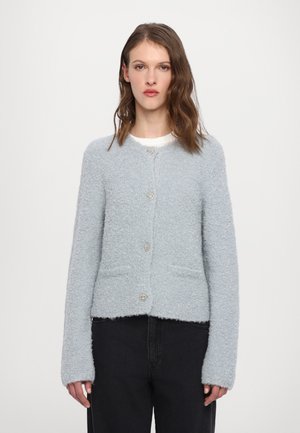 Cardigan moelleux bleu clair avec manches longues, encolure ronde, poches avant et boutons décoratifs. Le tissu a une apparence douce et texturée.