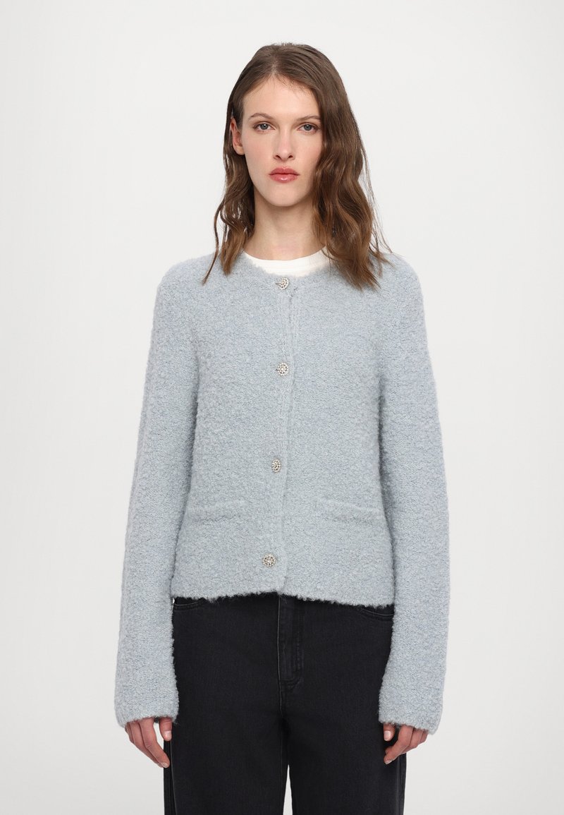 Lyseblå fluffy cardigan med lange ermer, rund hals, frontlommer og dekorative knapper. Stoffet har en myk, teksturert overflate.