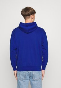 Felpa con cappuccio blu, con polsini e orlo a costine, in tessuto morbido e vestibilità rilassata, indossata con jeans chiari, vista da dietro.