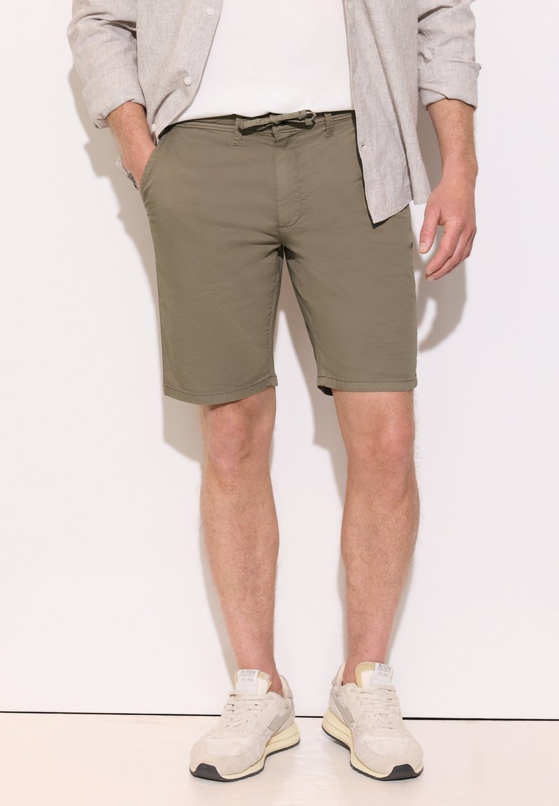 Homme portant un short vert olive, des baskets beiges, un t-shirt blanc et une chemise beige clair à boutons avec les manches retroussées. Mains dans les poches.