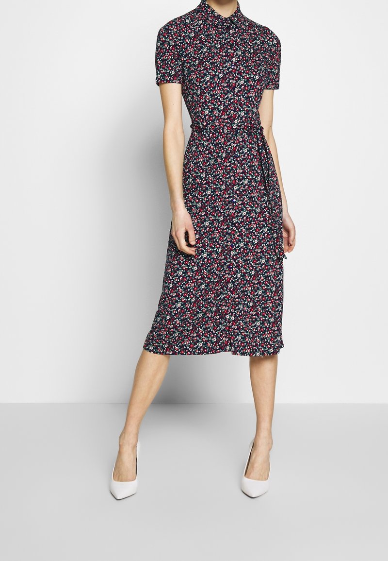 Kortärmad blommig klänning med en mörk marinblå bakgrund, prydd med små röda och vita blommor, en knapp-up krage och en bältesmidja.