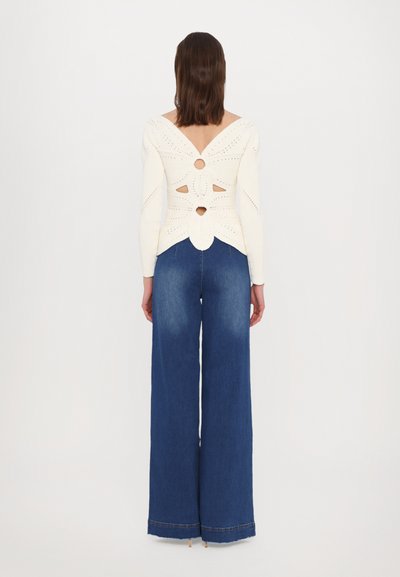 White knitted top with open back design featuring cutouts and a butterfly shape, paired with wide-leg blue denim jeans.