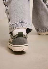 Zielono-szare sneakersy z wzorem kamuflażu, zamszowymi akcentami oraz białą podeszwą z detalem uśmiechniętej buzi na pięcie.