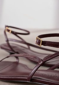 Bordeauxrote Ledersandalen mit schmalen Riemen, verziert mit einer goldenen Schnallendetails und flacher Sohle. Glatte Textur mit minimalistischem Design.