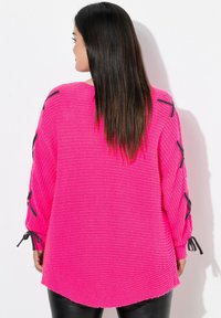 Frau mit glattem dunklem Haar, die einen hellrosa, locker gestrickten Pullover mit schwarzen Ribbons an den Ärmeln trägt, von einer weißen Wand abgewandt.