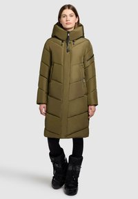 Kvinna som bär en olivgrön lång vadderad vinterjacka med huva och svarta snöboots, står mot en enfärgad ljus bakgrund.