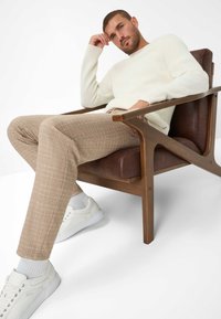 Een man zit op een houten stoel, gekleed in een gestructureerde witte trui en lichtbruine geruite broek, met witte sneakers en grijze sokken.