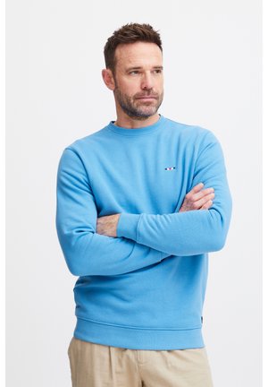 Light blue crewneck sweatshirt made of soft fabric, featuring a small emblem with red and white accents on the chest.