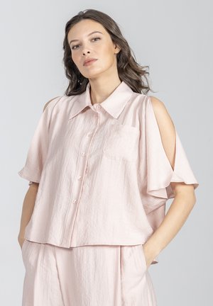 Kobieta ubrana w bladoróżową koszulę na guziki z wycięciami na ramionach i dopasowane luźne spodnie, stojąca z rękami w kieszeniach.