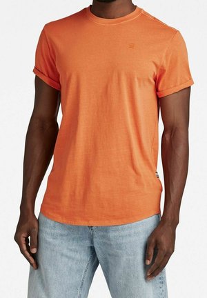 Oranžové bavlněné tričko s kulatým výstřihem, ohrnutými rukávy a malým logem na prsou. Hladký povrch, volný střih. Spárováno se světlými džíny.