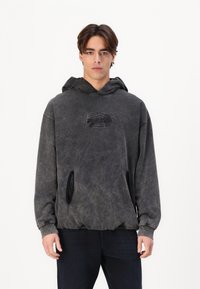 Sötétszürke kapucnis pulóver, amelyen elhalványult textúra látható, a mellkason logóval és fekete díszítéssel a elülső zsebein.