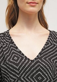 Černý top s designem do V, který je zdoben geometrickým bílým vzorem čtverců a diamantů. Hladká tkanina s mírným leskem.