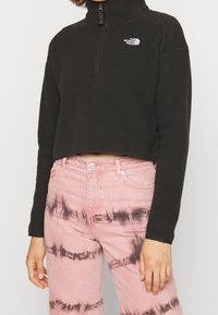 Persona che indossa un pullover in pile nero cropped con mezza zip e jeans rosa tie-dye con righe orizzontali nere.