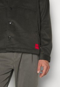 Homme portant une veste boutonnée gris foncé avec une étiquette rouge Hugo près de l’ourlet, et un pantalon plissé gris clair, main détendue le long du corps.