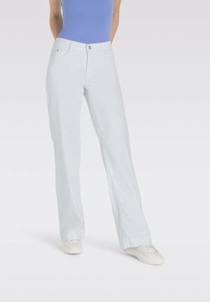 Person trägt weiße, gerade geschnittene Hose, weiße Sneakers und ein blaues, figurbetontes ärmelloses Oberteil und steht mit den Händen hinter dem Rücken.