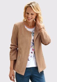 Beige faux-suède jas met een ronde halslijn, twee ritszakken aan de zijkant en driekwart mouwen, gecombineerd met een top met bloemenprint en een blauwe jeans.